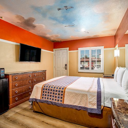 A cozy bedroom with a double bed, wall-mounted TV, dresser, microwave, fridge, red curtains, and a sky-themed ceiling mural, well-lit by windows.