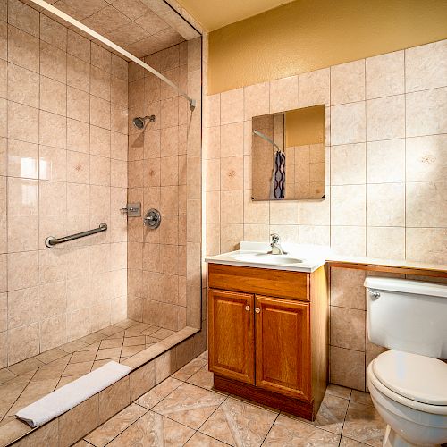 The image shows a bathroom with a walk-in shower, a vanity with a sink and mirror, and a toilet, all surrounded by beige tile walls and flooring.