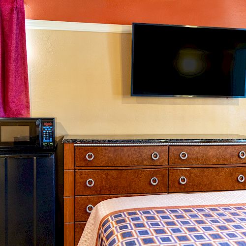 A room features a mounted TV, a dresser with drawers, a mini-fridge with a microwave on top, and a bed with a patterned bedspread in the foreground.