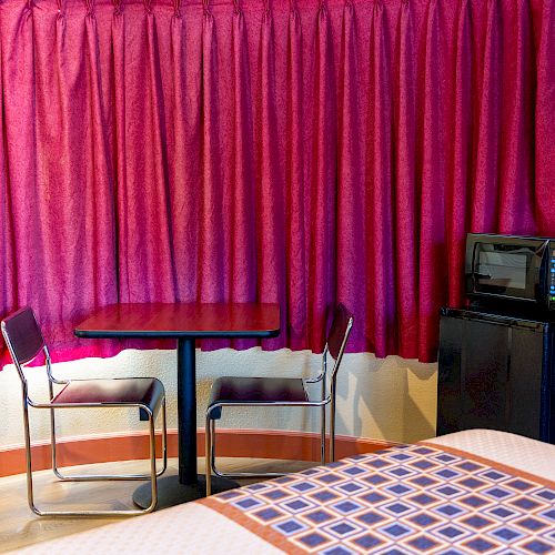 A hotel room features a table with two chairs, a bed, red curtains, a mini-fridge, and a microwave next to a small counter.