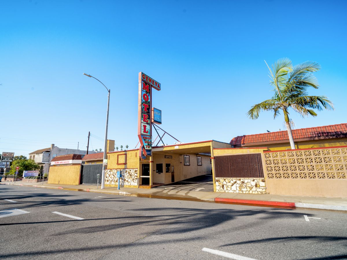 A vintage-style motel with a retro sign, palm trees, and a sunny sky in the background, located on an empty street corner.