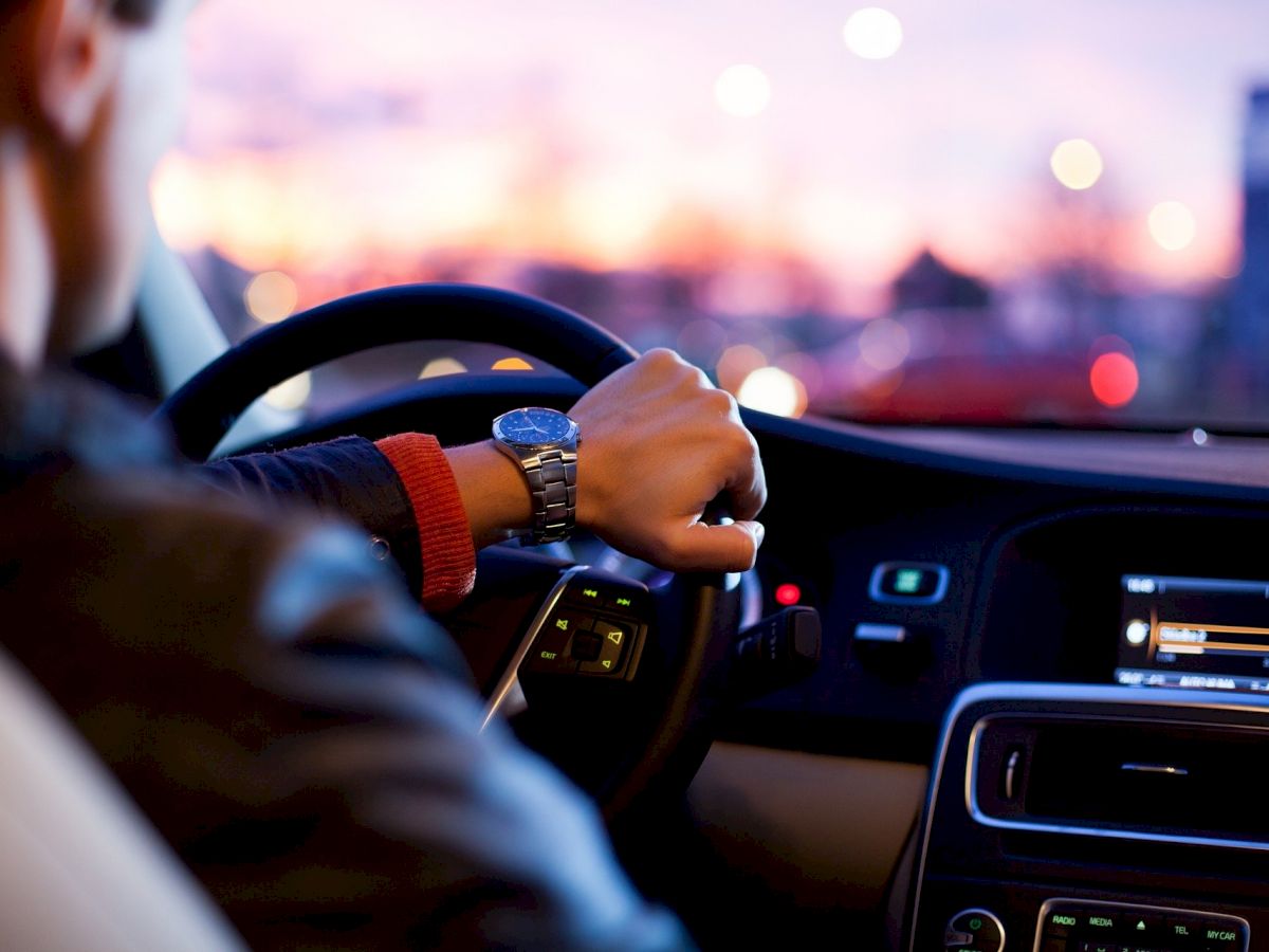 A person is driving a car at dusk, with their left hand on the steering wheel and the interior dashboard and controls visible.