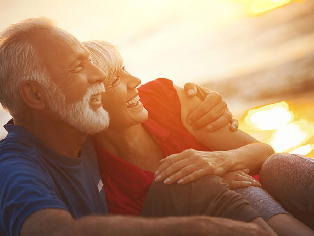 An older couple is sitting on a beach, smiling and embracing each other, with the sunset casting a warm glow in the background.