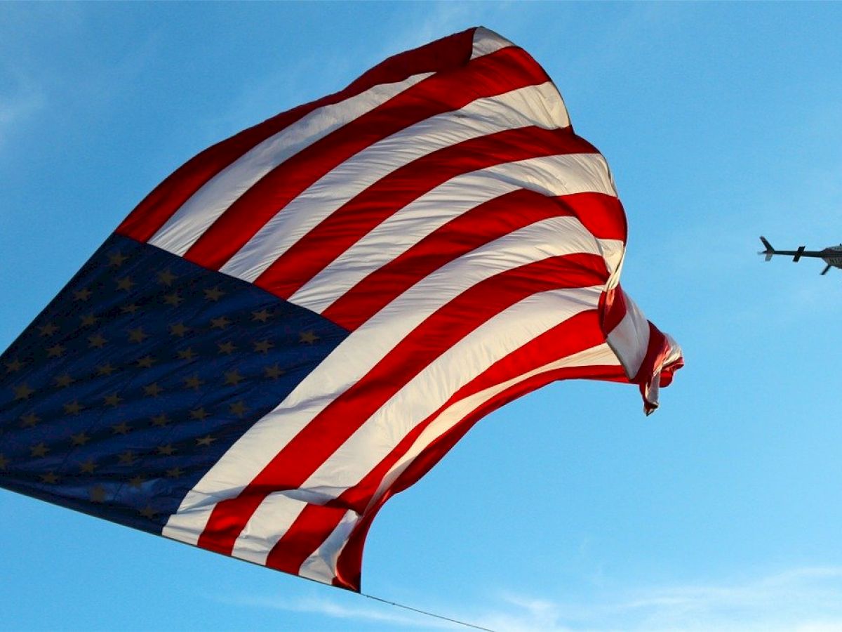 The image shows a large American flag waving in the sky with a helicopter flying nearby, set against a backdrop of blue sky and clouds.