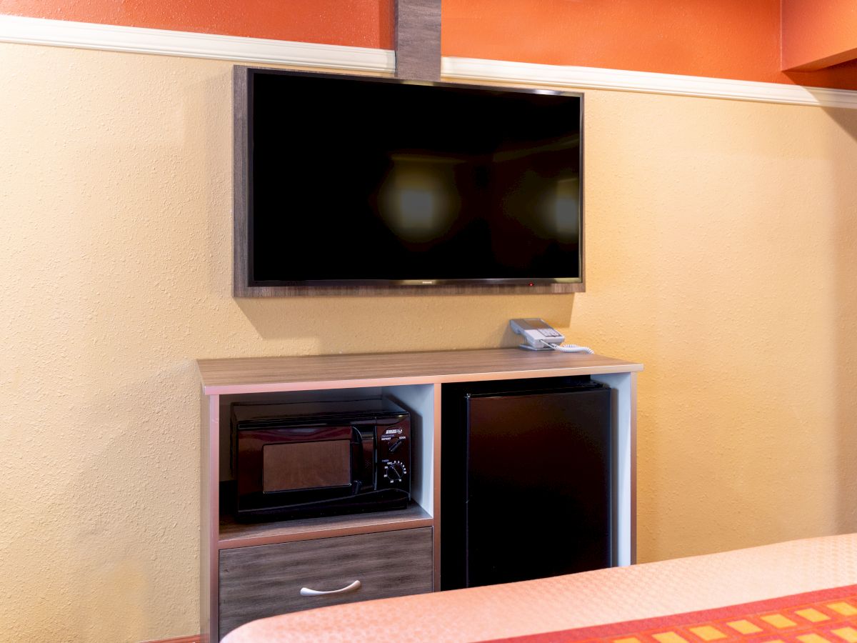 The image shows a mounted flat-screen TV above a small cabinet with a microwave and mini-fridge, next to a bed.