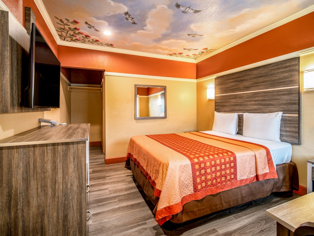 The image shows a hotel room with a king-size bed, wall-mounted TV, a dresser, wall lights, and decorated ceiling with painted clouds and birds.