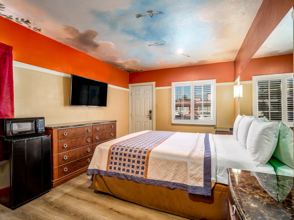 A cozy bedroom with a king-size bed, TV, microwave, dresser, and scenic ceiling mural, with vibrant red curtains and hardwood flooring.