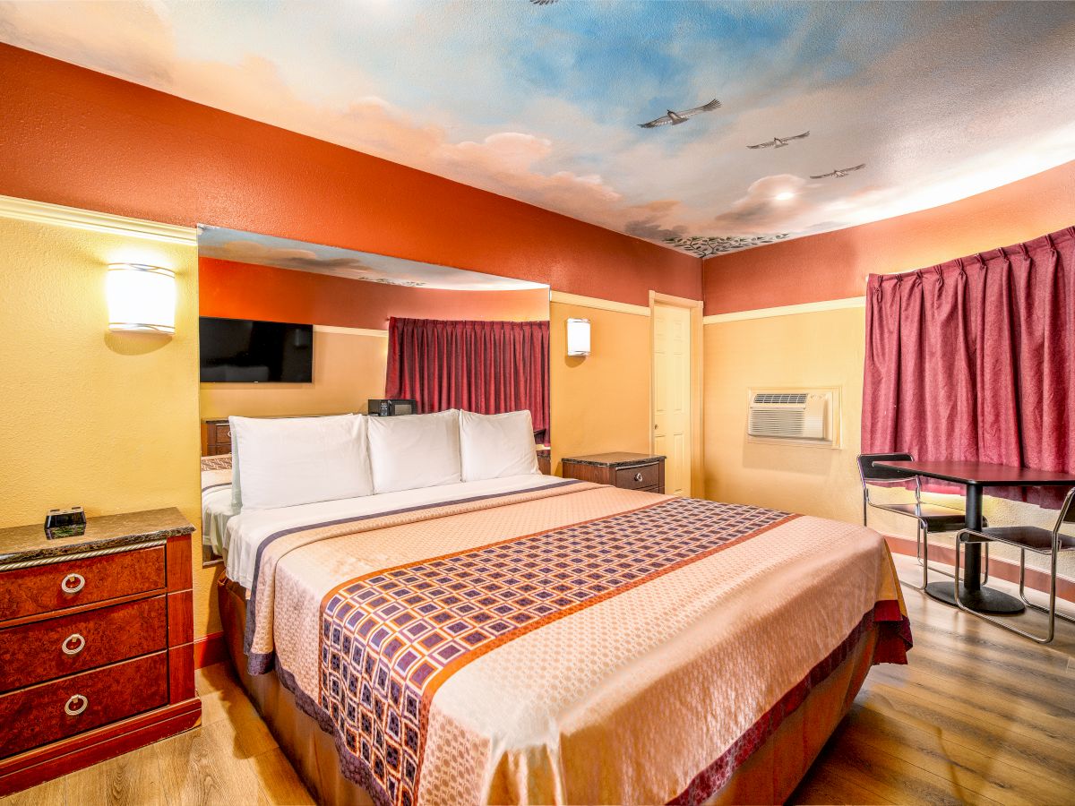 A hotel room with a large bed, vibrant colors, a painted ceiling, a dresser, TV, air conditioner, and a table with chairs by the window.