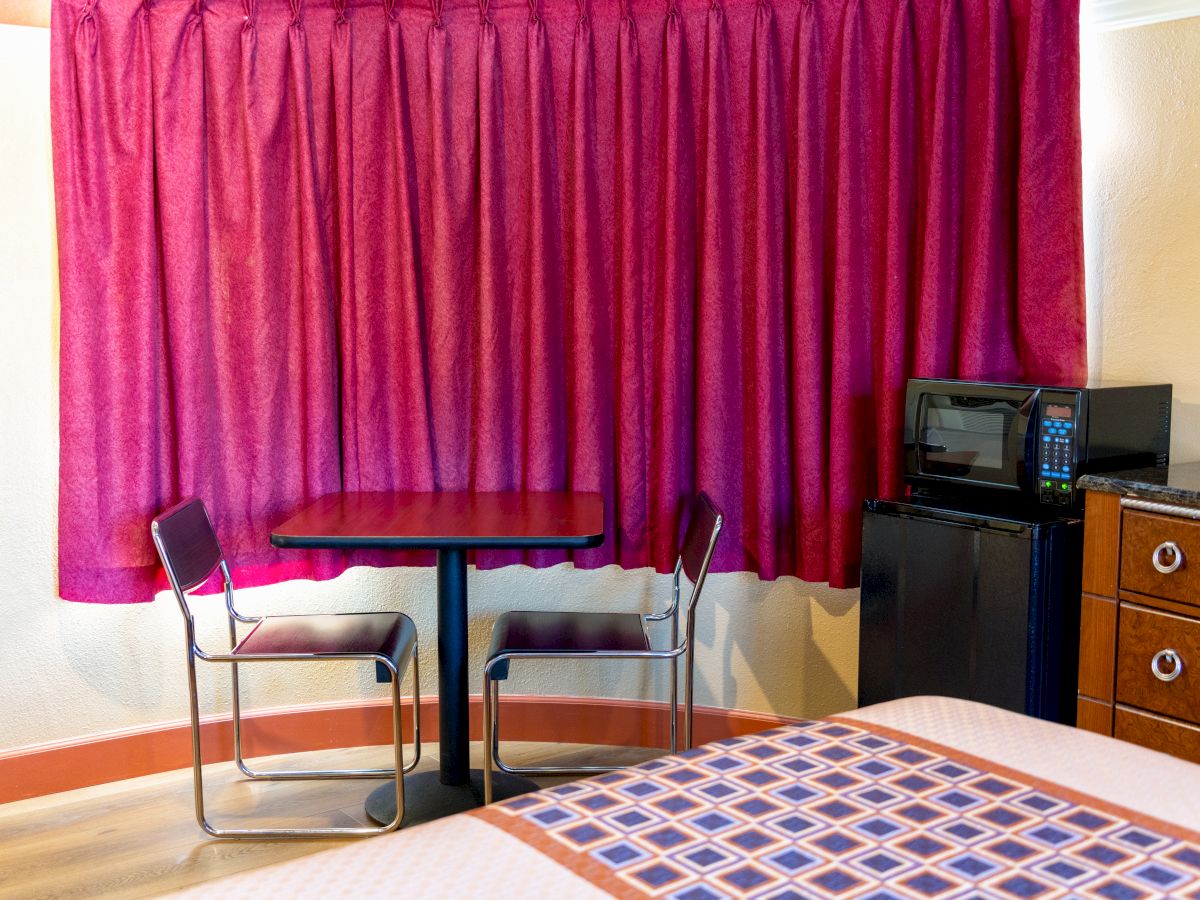 The image shows a room with magenta curtains, a table with two chairs, a microwave, a mini-fridge, and part of a bed.