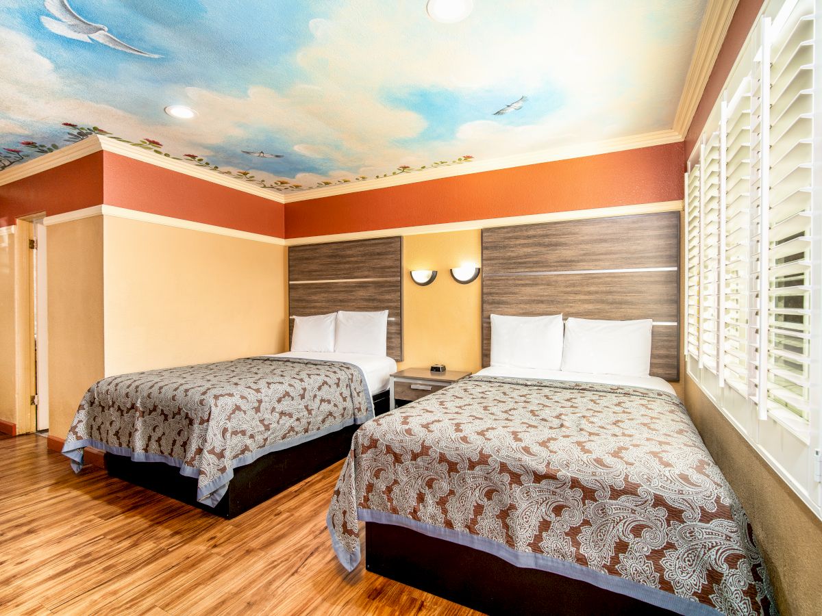 This image shows a hotel room with two double beds, wood flooring, and a painted ceiling depicting a sky with birds, featuring modern decor and lighting.