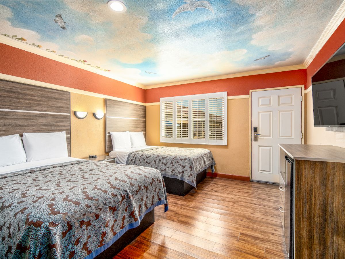 This image shows a hotel room with two beds, a TV on the wall, wooden flooring, and a ceiling painted with clouds and birds.