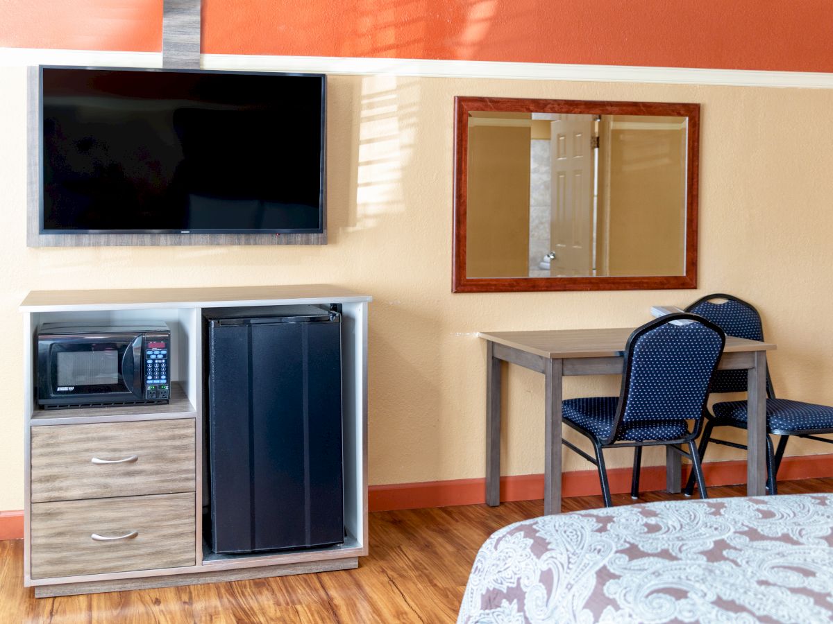 A room with a mounted TV, a cabinet with a microwave and fridge, a small table with two chairs, and a bed visible in the foreground.