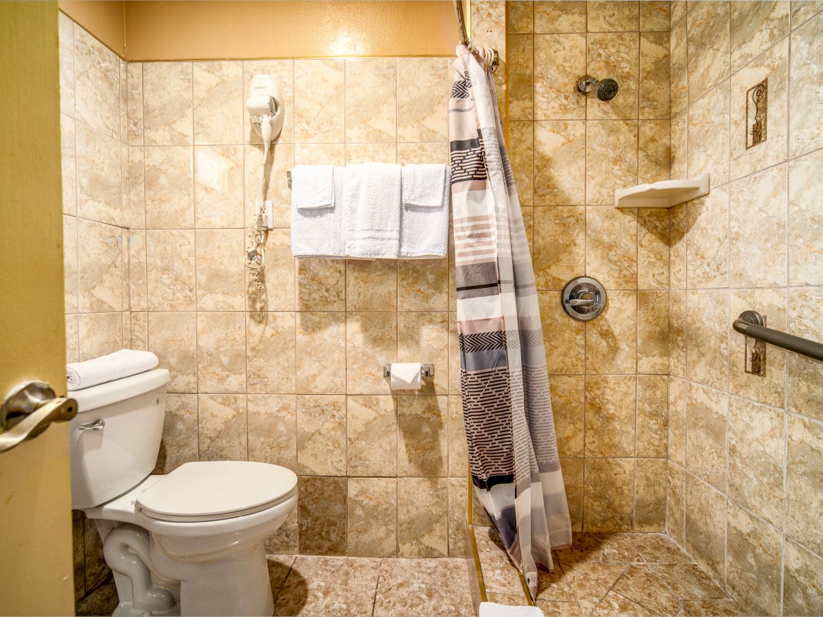 This image shows a bathroom with a tiled shower, toilet, towels on a rack, a hairdryer mounted on the wall, and a shower curtain.