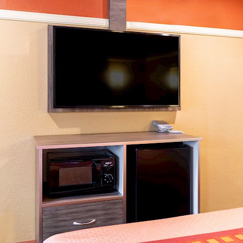 A wall-mounted TV is above a cabinet with a microwave and a mini-fridge, beside part of a bed with an orange and yellow checkered blanket.