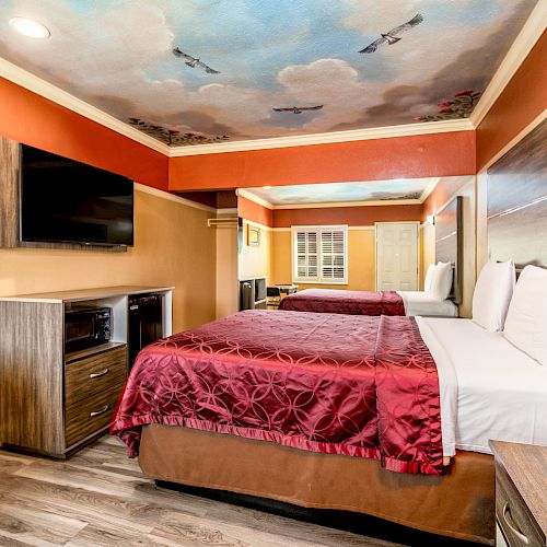 A hotel room with two beds, red bedspreads, a wall-mounted TV, and a painted ceiling with clouds and planes, designed with a warm color palette.