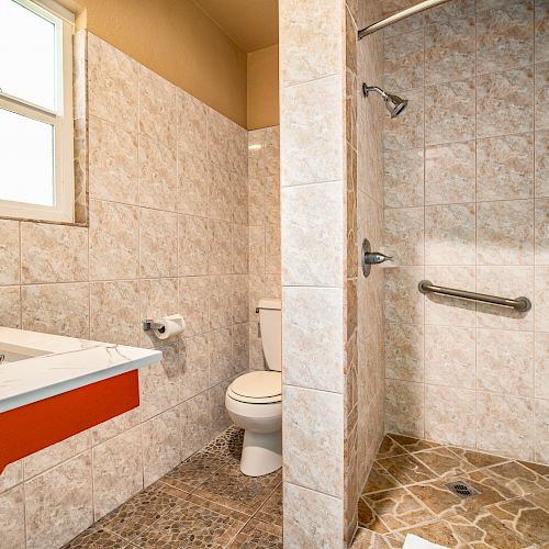 A bathroom with a sink, toilet, and walk-in shower featuring a grab bar, white tile walls, and a window providing natural light.