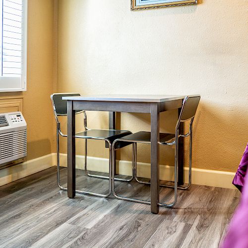 The image shows a small dining area with a table and two chairs, an air conditioner, and part of a bed with a red cover in a room.