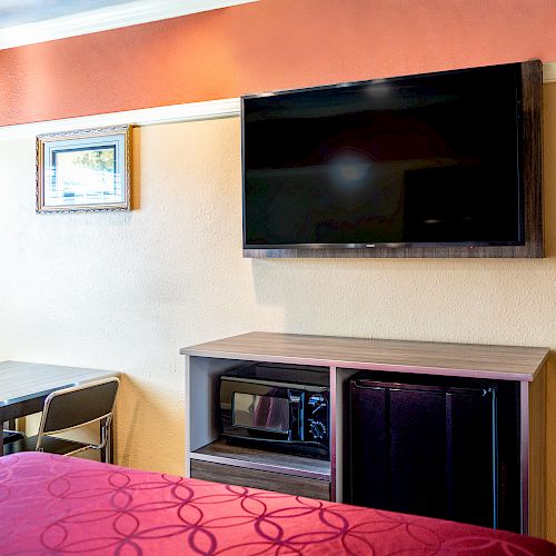 A hotel room wall with a mounted TV, a microwave, a mini-fridge, a small table with two chairs, a framed picture, and a bed with red bedding.