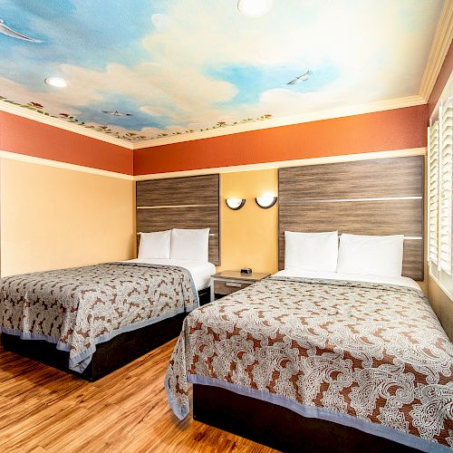 This image shows a hotel room with two double beds, a wooden floor, and a ceiling painted with a sky and clouds mural.