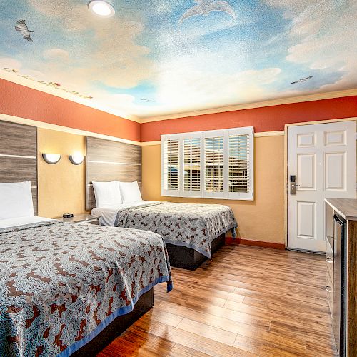 This image shows a well-lit hotel room with two beds, a TV, and a sky-themed ceiling mural. The room has wooden flooring and modern furnishings.