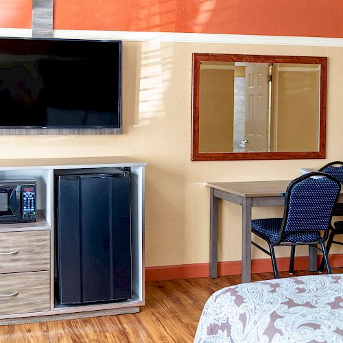 A hotel room features a wall-mounted TV, mirror, table with two chairs, microwave, and mini-fridge on a wooden dresser, completed by a bed in the foreground.