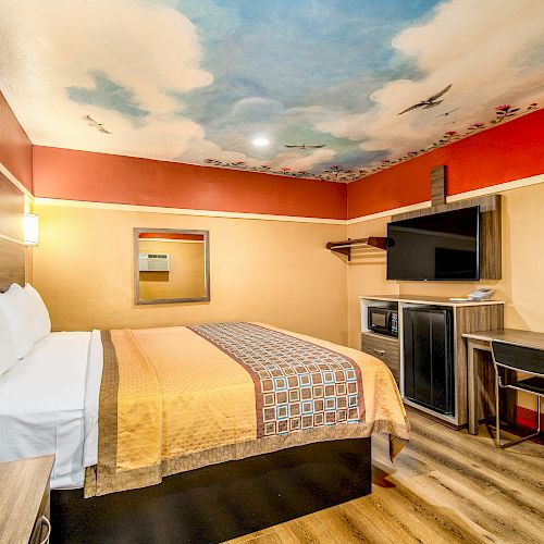 A cozy hotel room with a mural ceiling, bed, TV, refrigerator, desk, and chair. The walls are painted in warm tones, adding a snug ambiance.