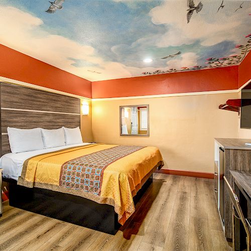 A cozy hotel room with a king-sized bed, a colorful ceiling mural, a wall-mounted TV, a desk, nightstands, and a mini-fridge.