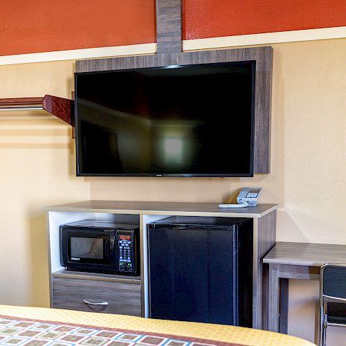 The image shows a hotel room setup with a mounted TV, microwave, mini-fridge, small desk, and a chair against a light-colored wall.