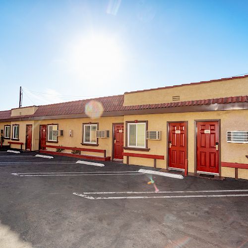 A small motel with several rooms, each with a red door and window unit AC, and an empty parking lot under a bright, sunny sky.