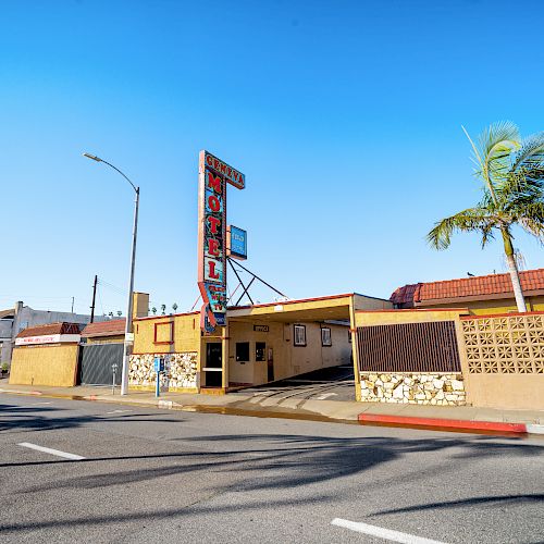 A small roadside motel with a neon sign, palm tree, and mid-century architecture, located on a sunny street with minimal traffic.