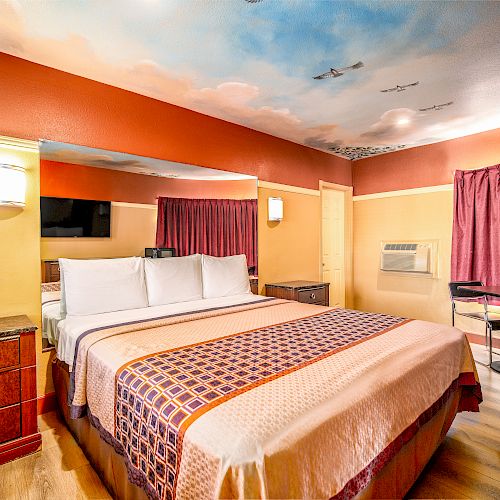 A neatly made bed in a cozy hotel room featuring warm colors, a painted ceiling, a nightstand, a TV, and a small dining area next to curtained windows.