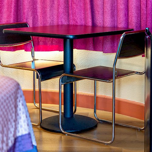 A small table with two chairs is set near a pink curtain and adjacent to a bed with a pink blanket in a cozy room.