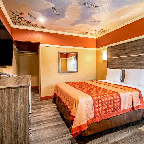 A hotel room with a double bed, wall-mounted TV, dresser, and a painted ceiling featuring birds and clouds. Warm tones and modern furnishings complete the look.