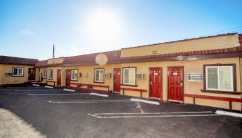 The image shows a bright sunny day over a single-story motel with red doors and parking spaces in front of the rooms.