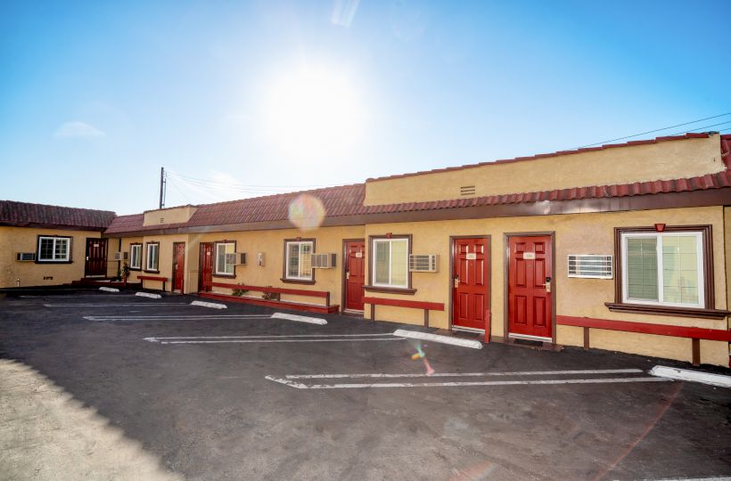 The image shows a bright sunny day over a single-story motel with red doors and parking spaces in front of the rooms.