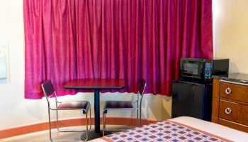 A room with pink curtains, table, two chairs, microwave, small fridge, and part of a bed with a patterned cover is visible, ending the sentence.