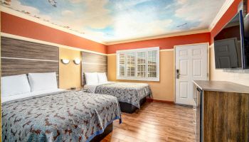 A hotel room with two double beds, wood flooring, a wall-mounted TV, and a painted sky ceiling with clouds and birds.