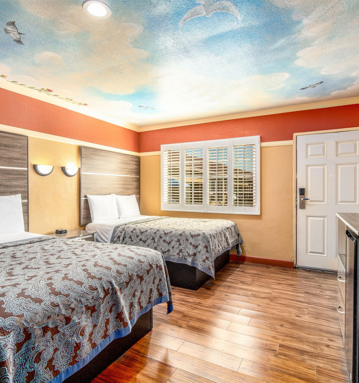 This image shows a hotel room with two beds, a wall-mounted TV, wooden flooring, and a painted sky-themed ceiling. The room looks clean and well-lit.