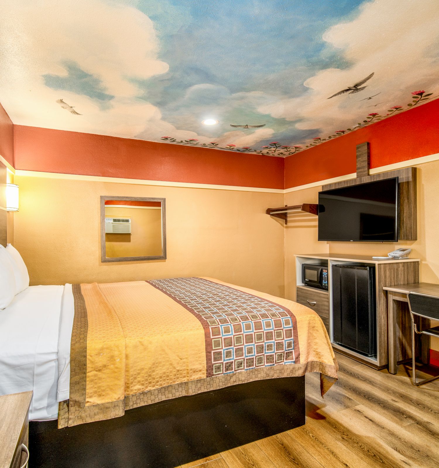 The image depicts a hotel room featuring a bed, desk, wall-mounted TV, mini-fridge, and a painted ceiling with a sky theme, creating a calm ambiance.