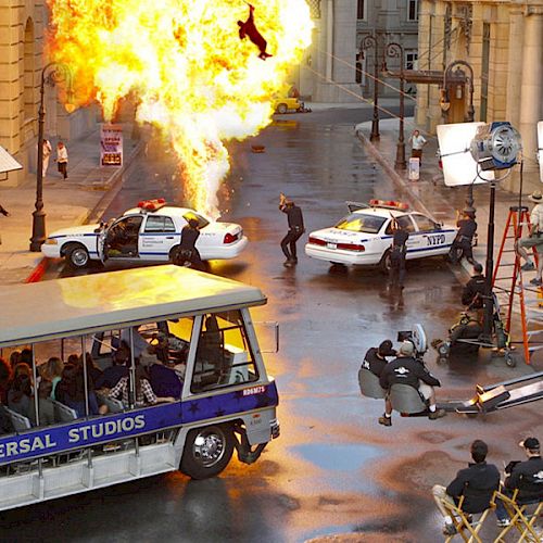 A Universal Studios tour passes by a film set featuring a staged car chase and explosion, capturing onlookers' attention with the dramatic scene.