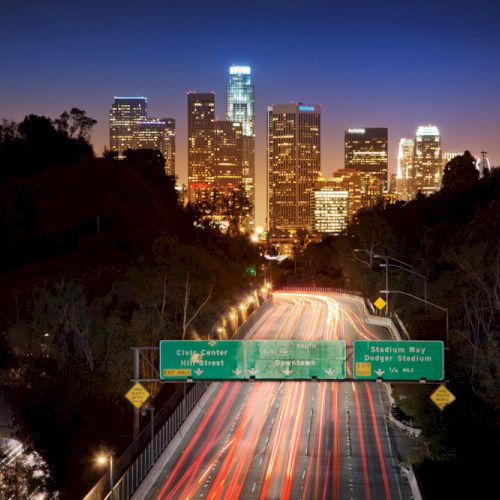 Nighttime cityscape with skyscrapers in the background and light trails from cars on a freeway in the foreground; green road signs visible.