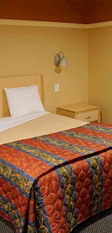 A cozy room with a bed featuring a colorful quilt, two side tables with lamps, a framed picture on a yellow wall, and a chair in the corner.